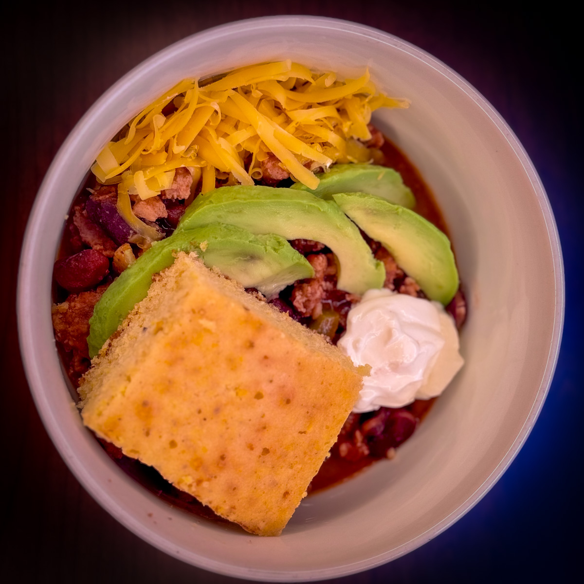 A bowl of turkey chili with shredded cheese, sliced avocado, a dollop of sour cream, and a slice of cornbread.